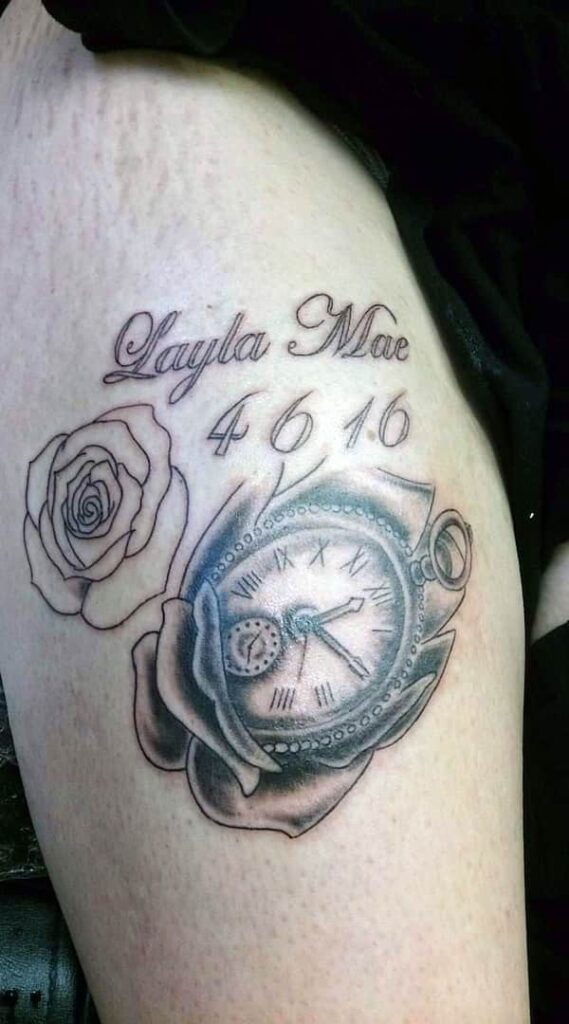 Tattoo of a clock in a rose, with accompanying name and date, on upper arm. John at Rogsy Tattoo Studio.